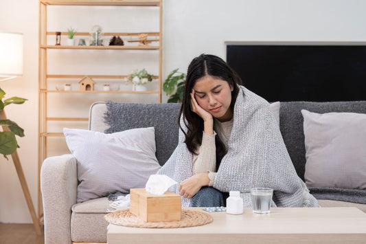 Woman resting on a sofa with a blanket, tissues, medicine, and water while feeling unwell