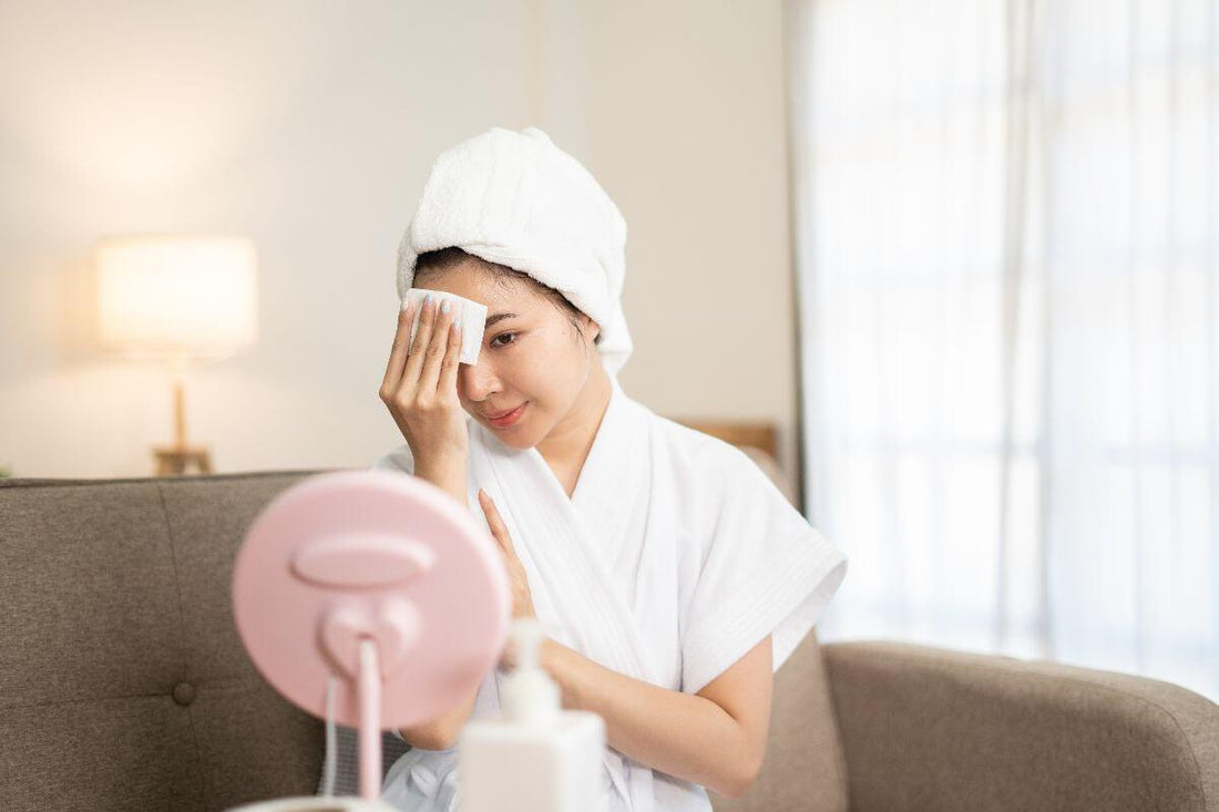 Woman in a bathrobe and towel gently wiping her face while looking into a tabletop mirror