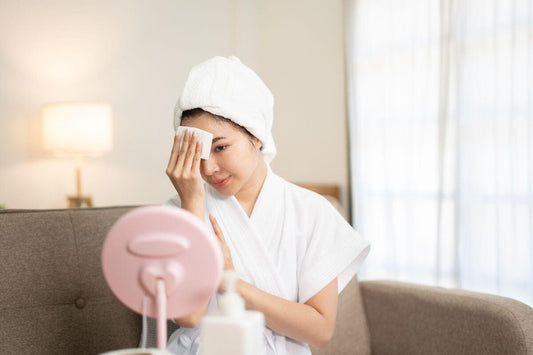 Woman in a bathrobe and towel gently wiping her face while looking into a tabletop mirror