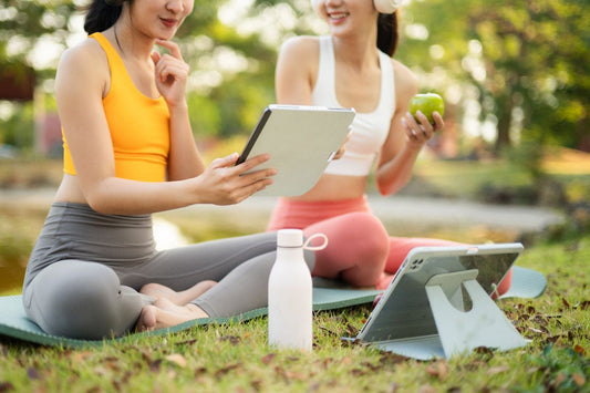 Two women in workout clothes sitting outdoors with tablets and a water bottle while planning their wellness routine